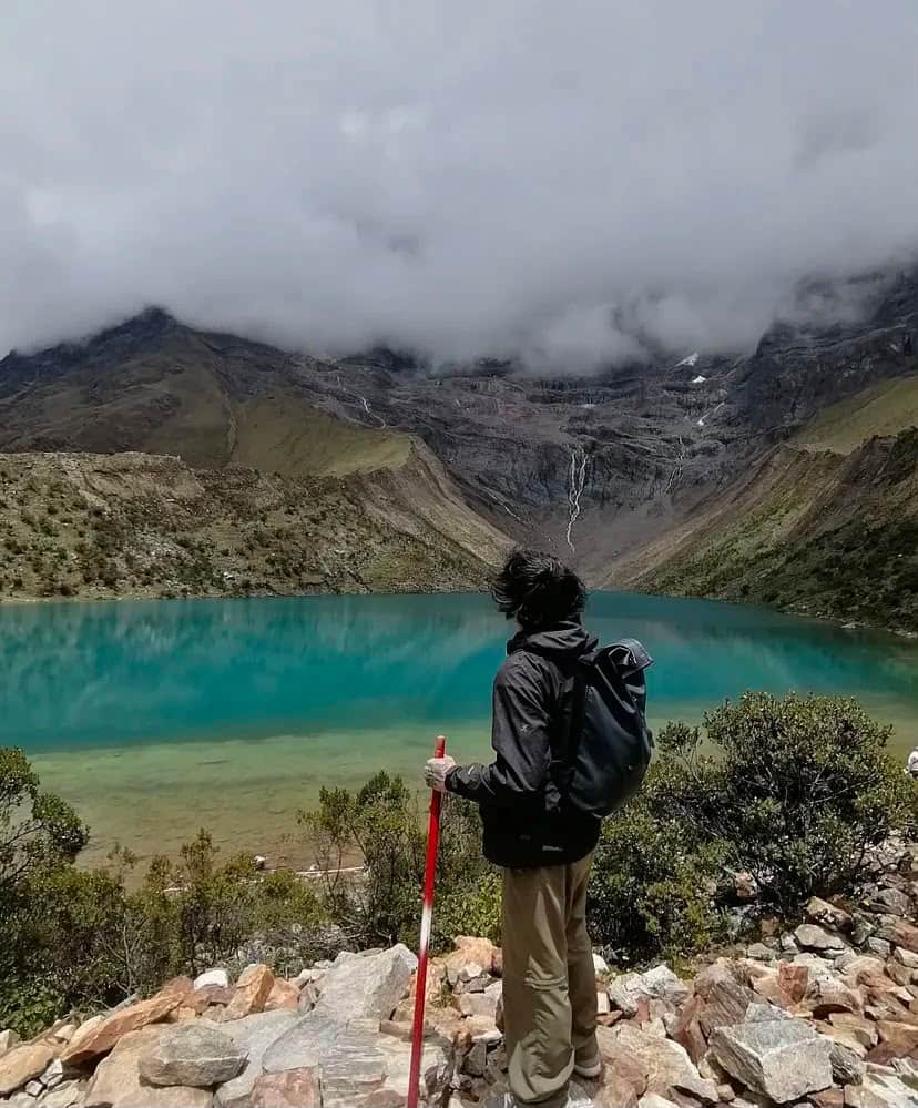 Traveller with a trekking pole standing on the rocky shore of Laguna Humantay, looking over the lake and glacier face.
