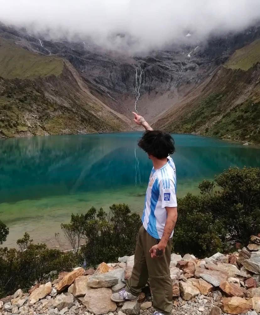 Traveller in an Argentina national football jersey pointing up at the waterfall flowing from the glacier face, looking over the turquoise Laguna Humantay.