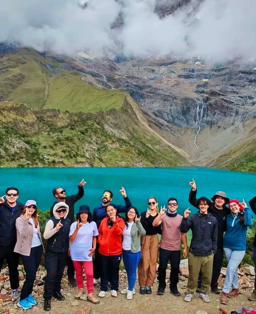 Group of thirteen travellers posing together at the rocky shore of Laguna Humantay, with the glacier-fed turquoise lake and snow-capped peak above them.