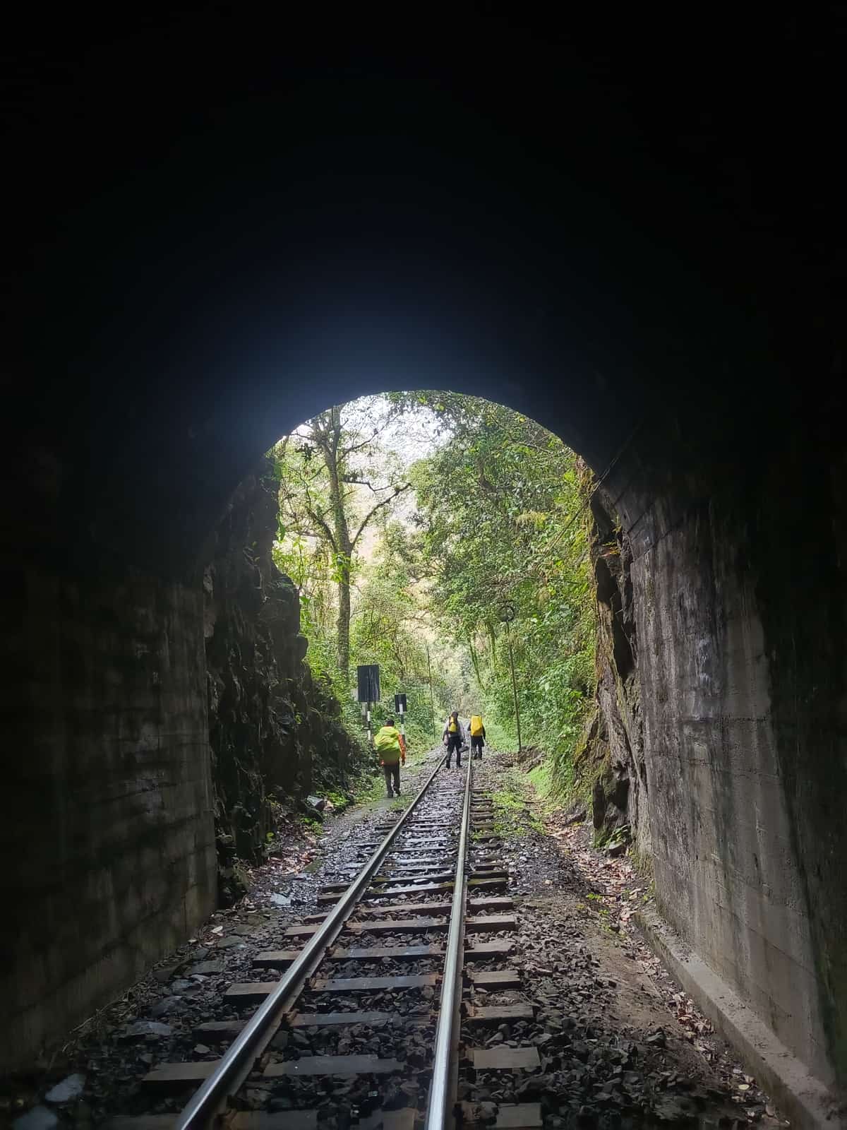 Three trekkers with backpacks walking out of a railway tunnel toward the cloud forest, on the train route to Aguas Calientes.
