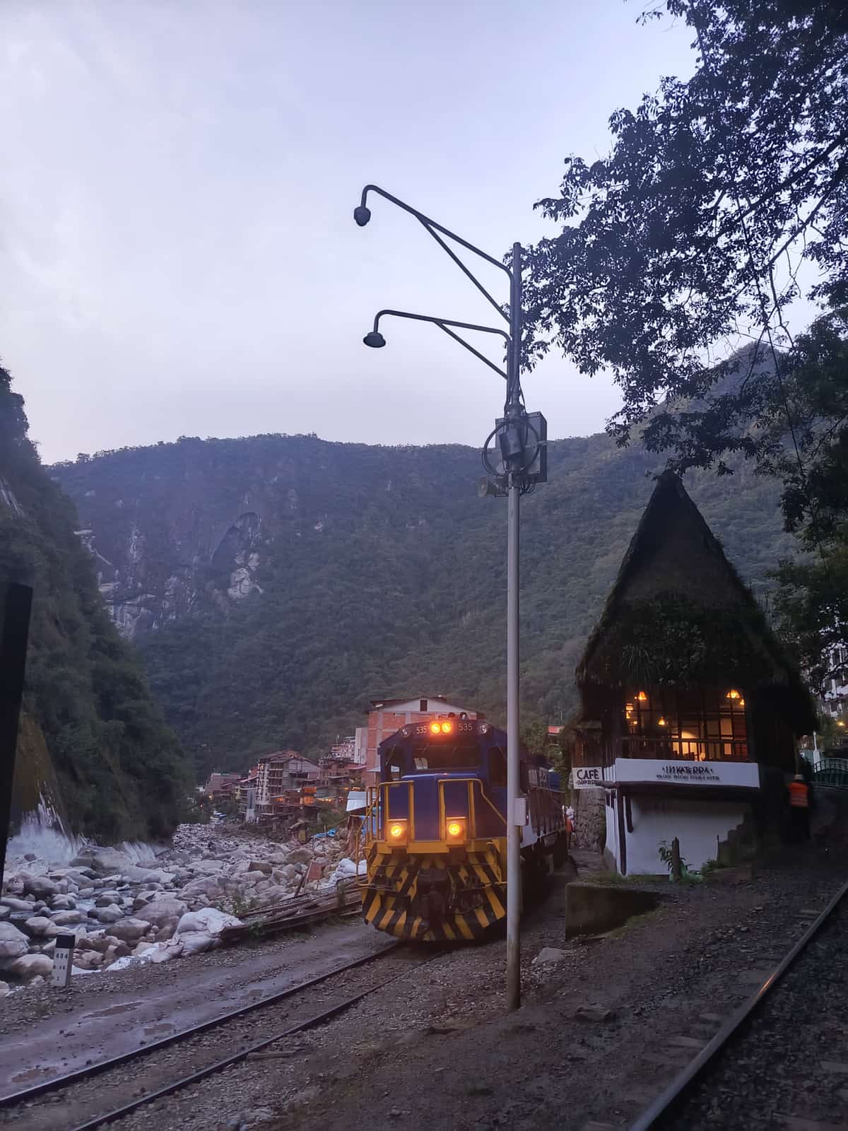 Train locomotive number 535 arriving at Aguas Calientes station at dusk, with the Café Inkaterra building on the platform and Andean mountains behind.
