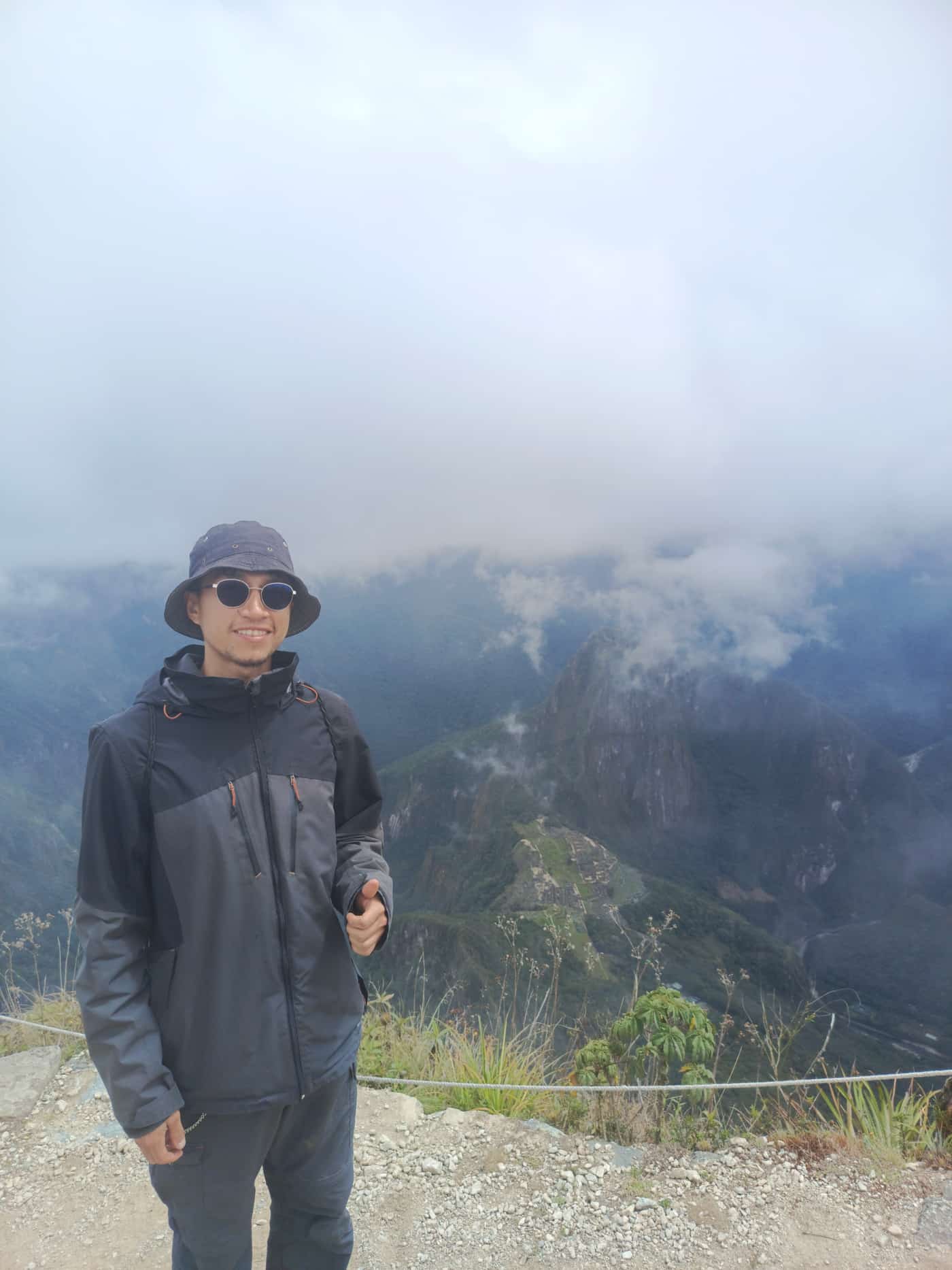 Mountain guide smiling at the Wayna Picchu summit, with the Machu Picchu citadel and the Wayna Picchu peak rising through clouds behind him.
