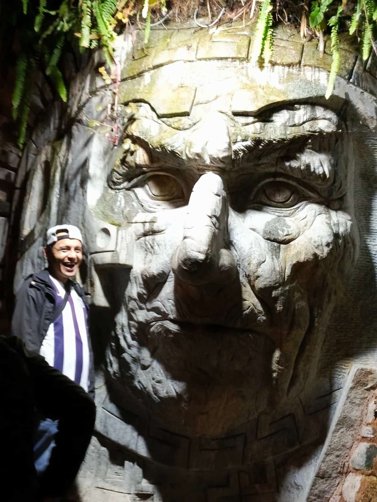 Traveller in a dark jacket laughing and posing in front of a giant Inca stone-face carving inside the Machu Picchu citadel area.