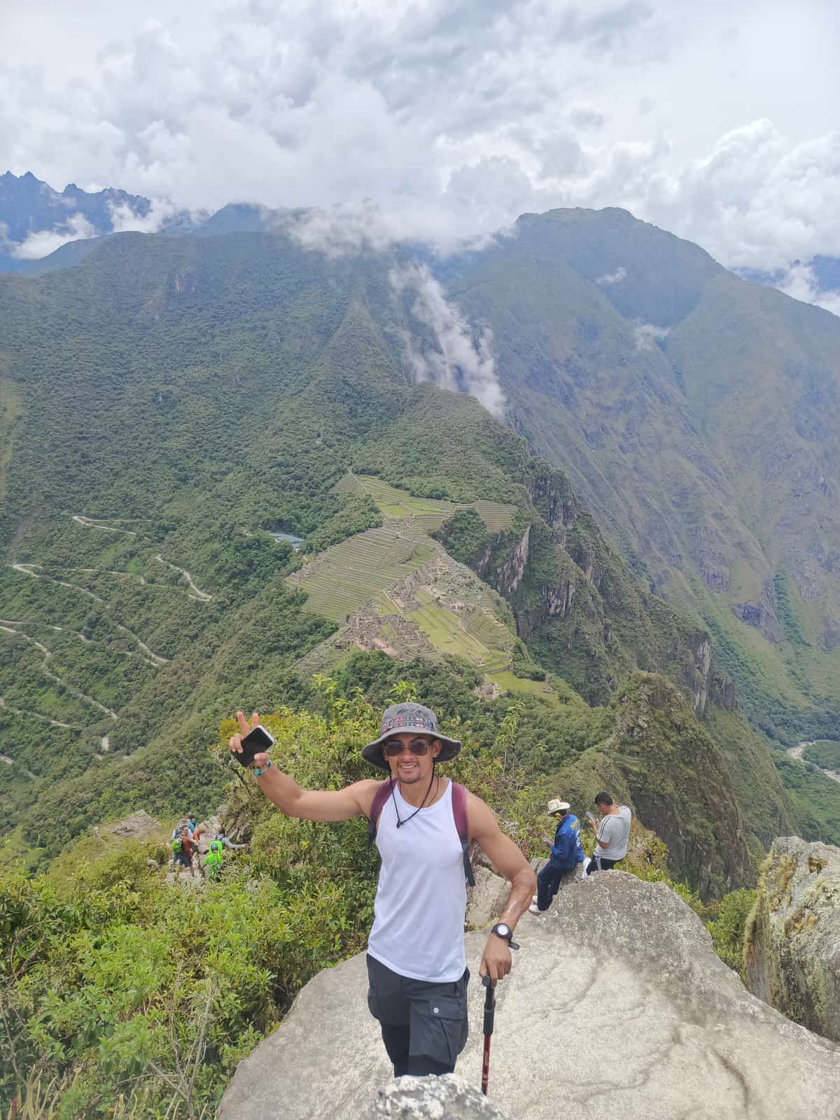 Traveller in a white tank top and dark hat giving a peace sign at the Wayna Picchu summit, with the full Machu Picchu citadel visible far below and other climbers nearby.