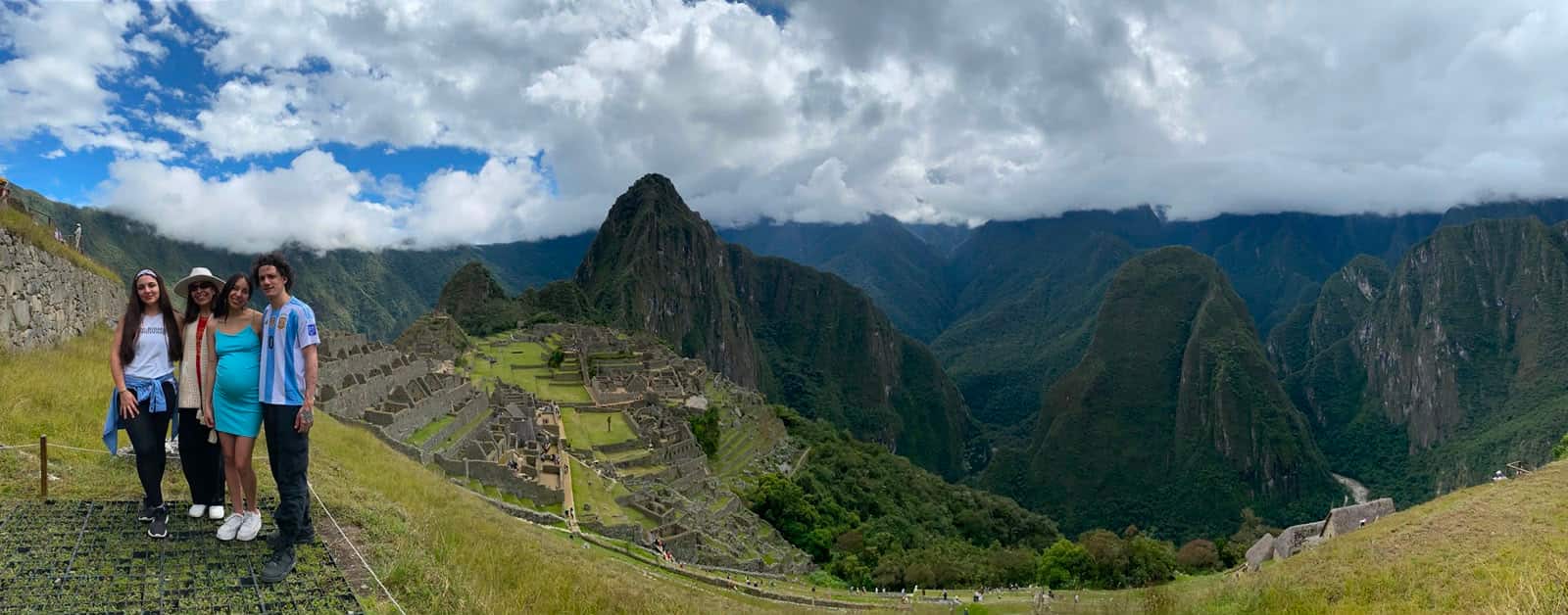 Group of four travellers at the iconic Machu Picchu citadel viewpoint — three women and a man, including one in an Argentina jersey — with the full citadel terraces and the Wayna Picchu peak spread out behind them.
