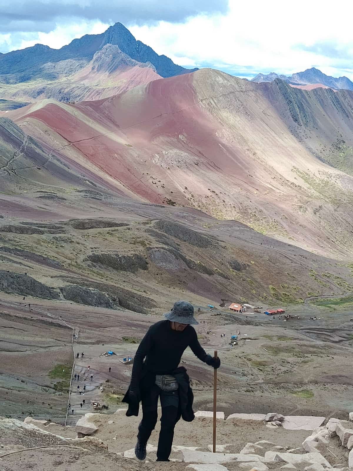 Mountain guide descending from Vinicunca's red slopes with a walking stick; the multi-coloured rainbow mountain behind him, and dozens of tiny climbers still hiking the winding trail below.