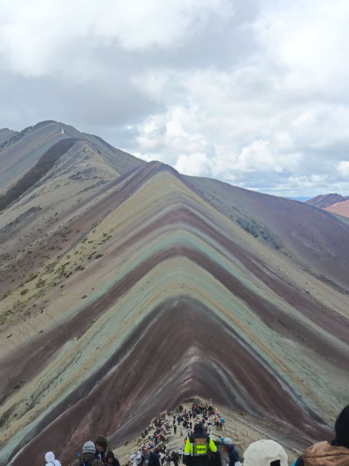Crowd of climbers with colourful gear near the summit of Vinicunca (Rainbow Mountain), with a yellow-vest security guard reading 'SEGURIDAD VINCUNCA' and the mountain's banded mineral striations visible.
