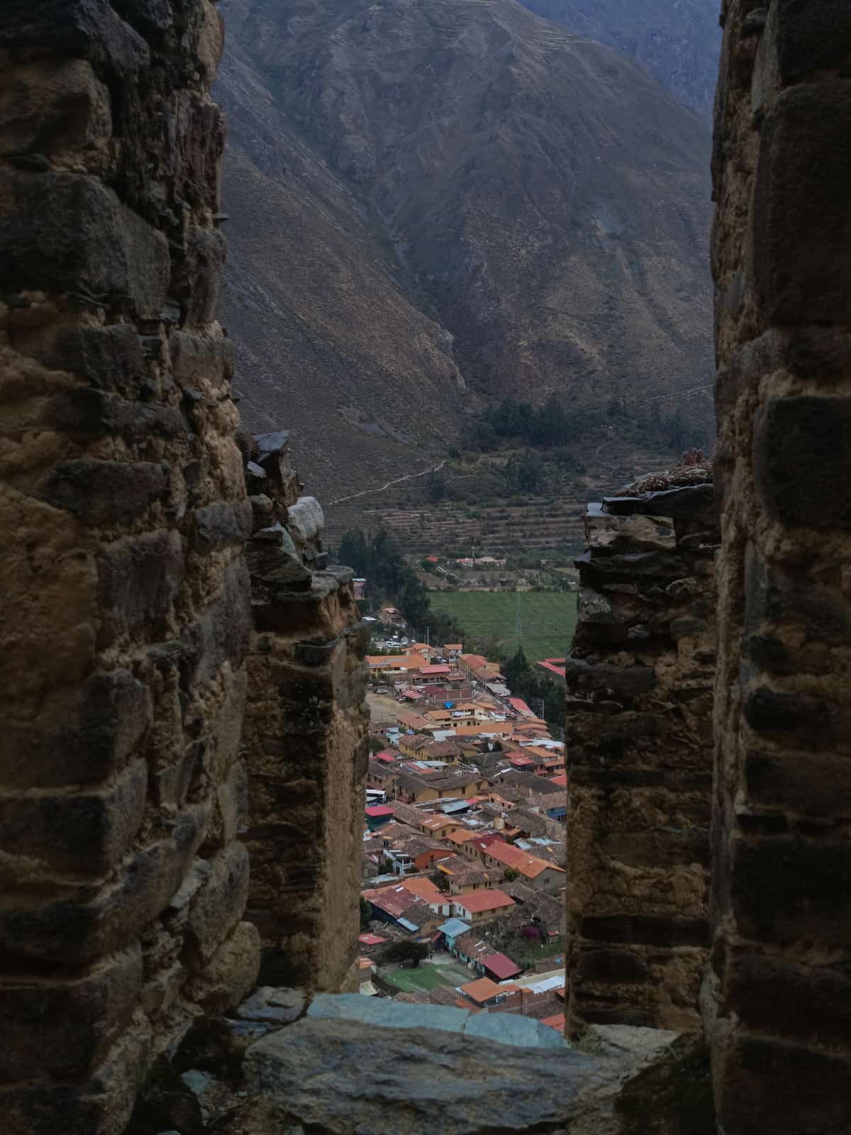View through an Inca-stone slit-window down at the red-roofed town of Ollantaytambo, the terraced Sacred Valley floor, and a steep Andean mountainside.