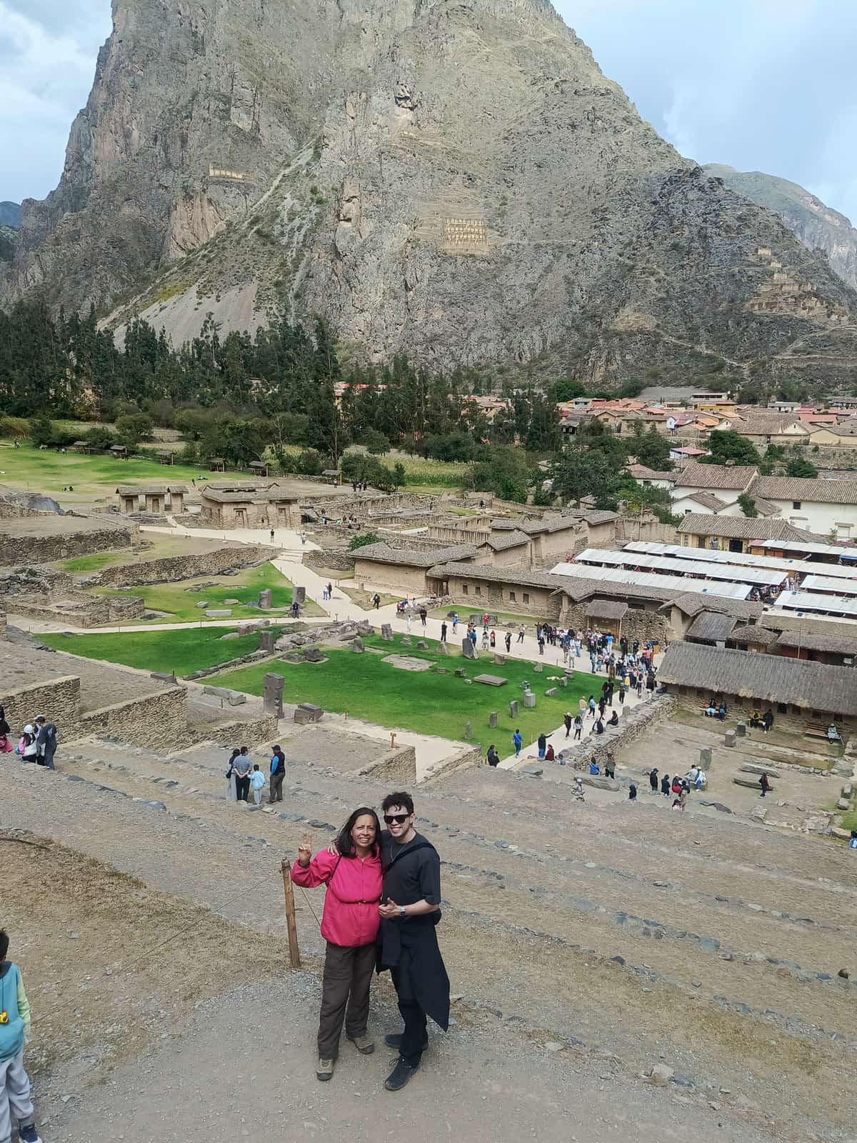 Two travellers posing with smiles at Ollantaytambo, with the terraced Inca storehouses and stone steps behind them and other tourists exploring the site.