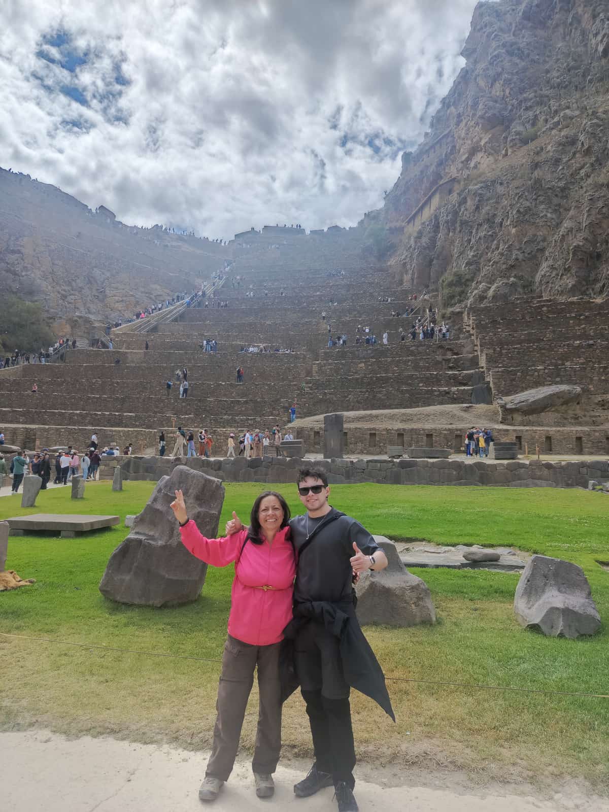 Two travellers giving thumbs-up poses at Ollantaytambo, smiling broadly with the long staircase of Inca terraces climbing the cliff face behind them and a chain of tiny tourists ascending.